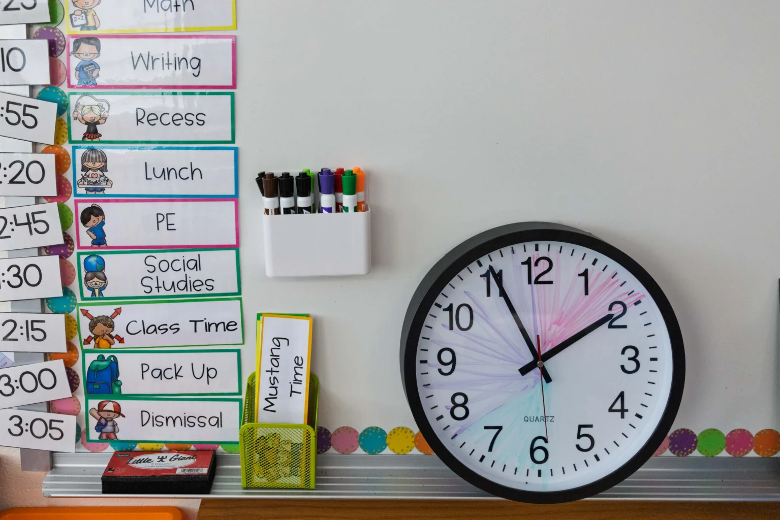 A classroom schedule is posted on a whiteboard next to a wall clock showing 1:50, with markers and a yellow note labeled "Mustang Time" nearby.