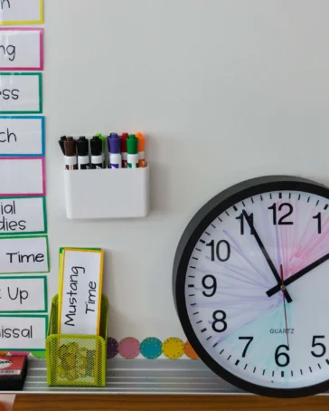 A classroom schedule is posted on a whiteboard next to a wall clock showing 1:50, with markers and a yellow note labeled "Mustang Time" nearby.