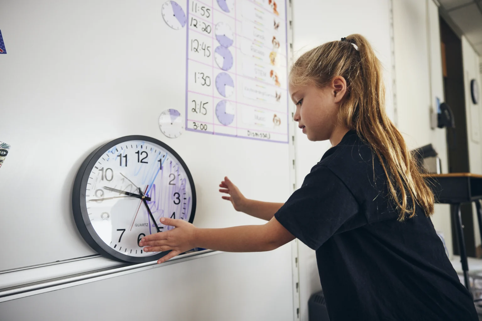 A young girl adjusts a wall clock in a classroom, with a daily schedule and time-related posters visible in the background.