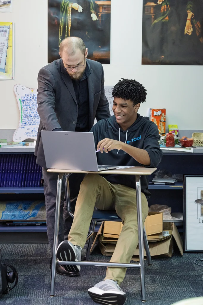 A teacher stands beside a seated student, both looking at a laptop on the student's desk in a classroom with books and posters in the background.