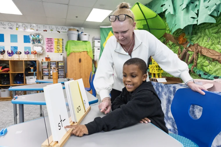 A teacher helps a student read phonics cards at a desk in a colorful, decorated classroom.