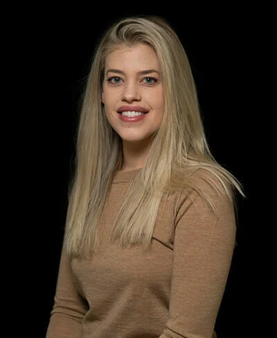 A woman with long blonde hair wearing a brown sweater smiles at the camera against a black background.
