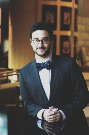 A man wearing glasses, a dark suit, and a bow tie stands indoors with his hands clasped, facing the camera. Framed artwork and warm lighting are visible in the background.