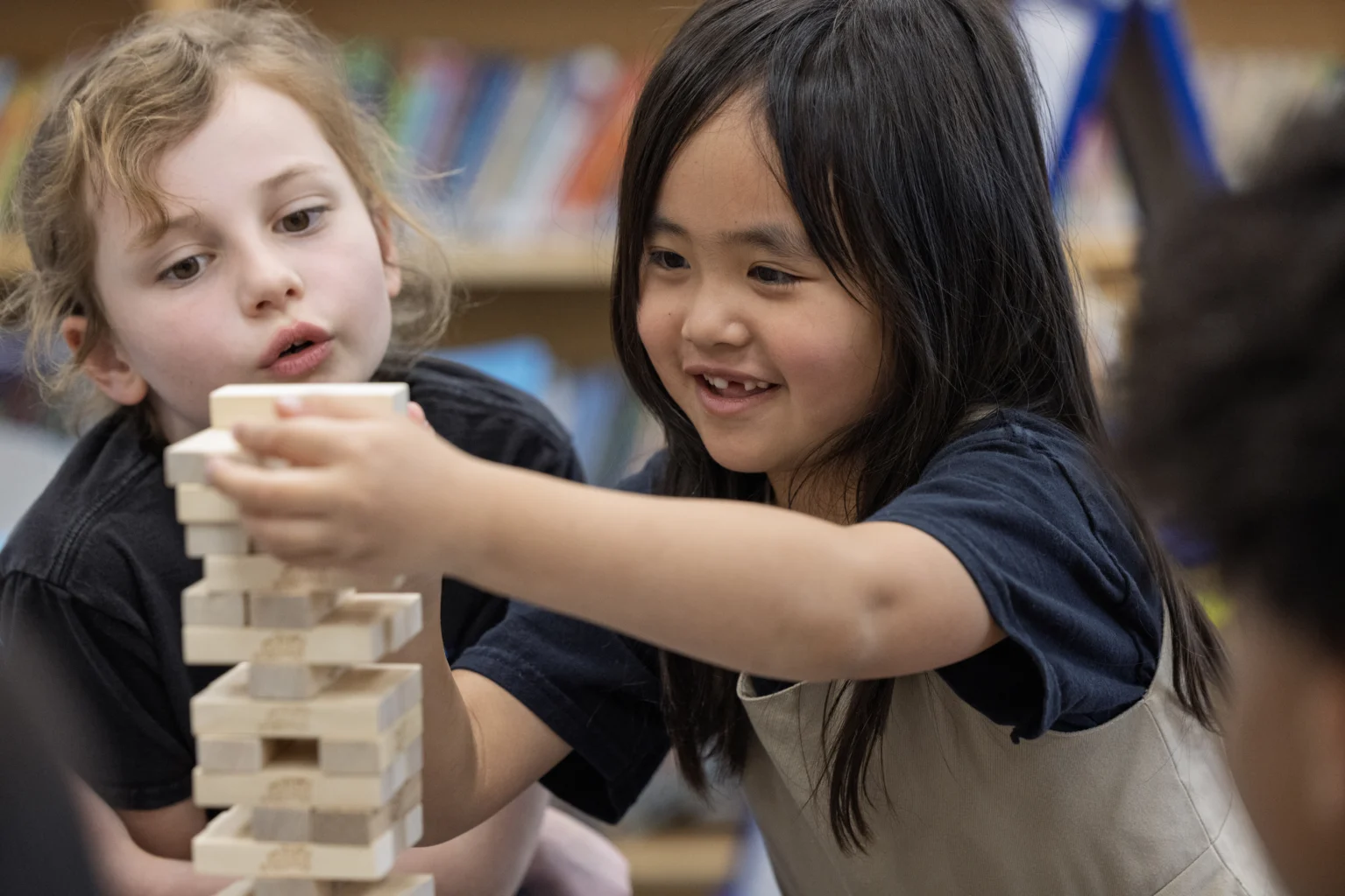 Two young children focus on carefully stacking wooden blocks while playing a game together in a classroom setting.