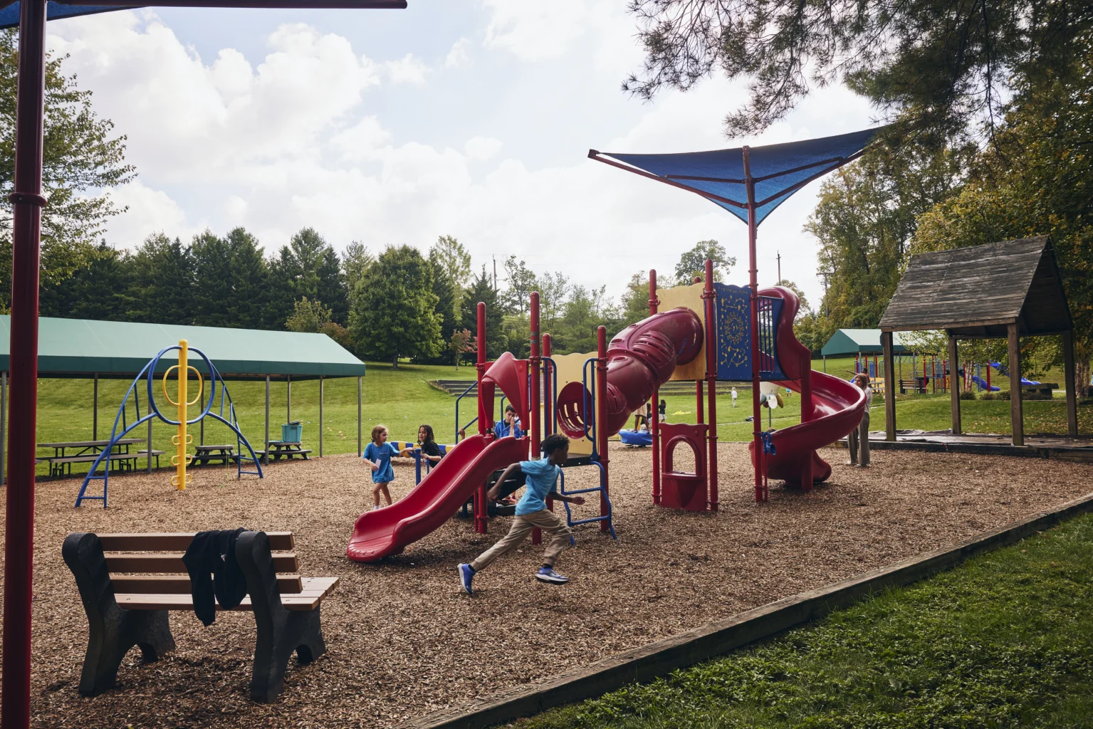Children play on a red playground structure with slides and tunnels in a park. Benches, a climbing area, and trees are visible in the background on a sunny day.