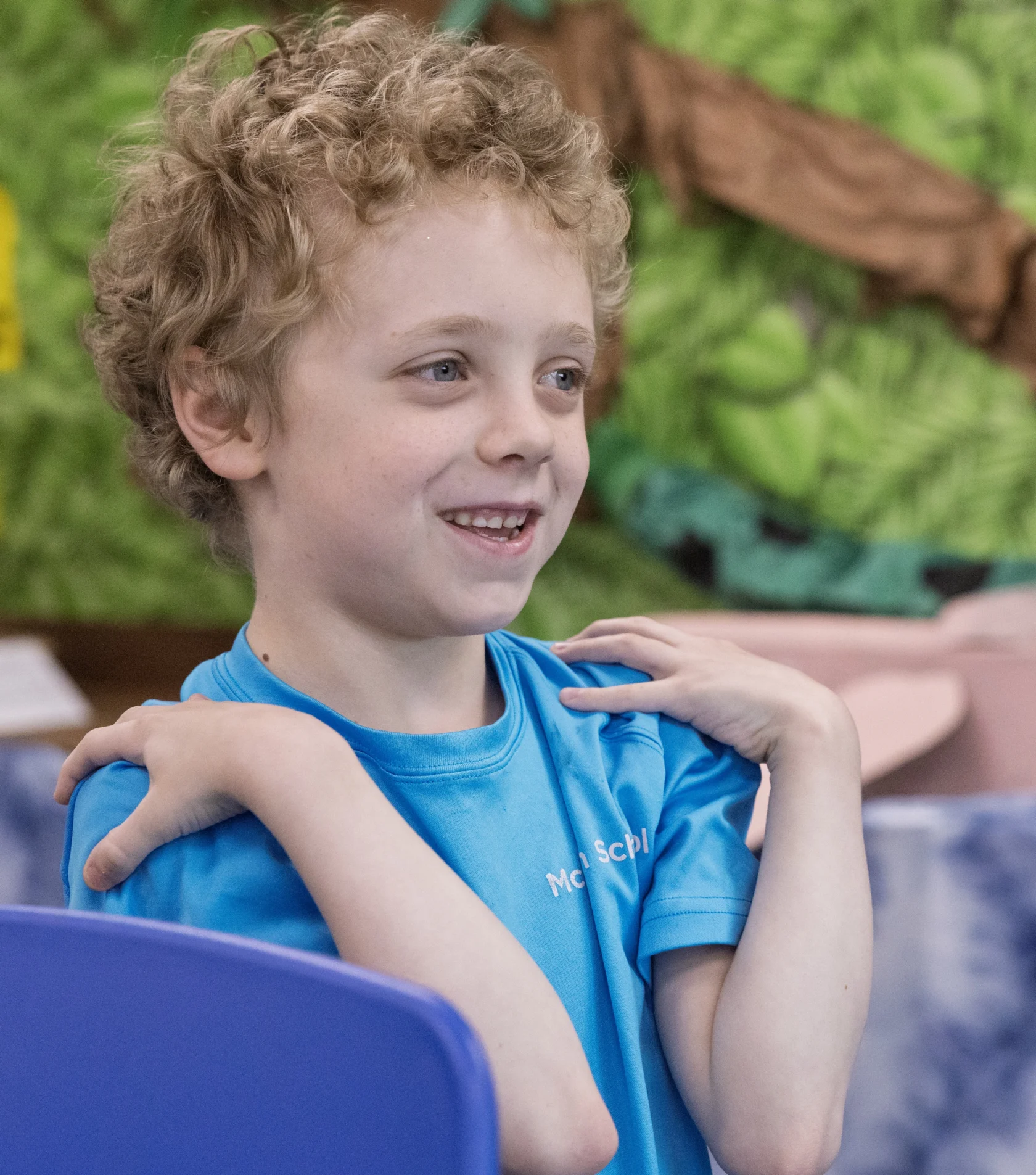 A young child with curly hair in a blue shirt smiles and crosses their arms over their chest, sitting indoors with a green and brown background.