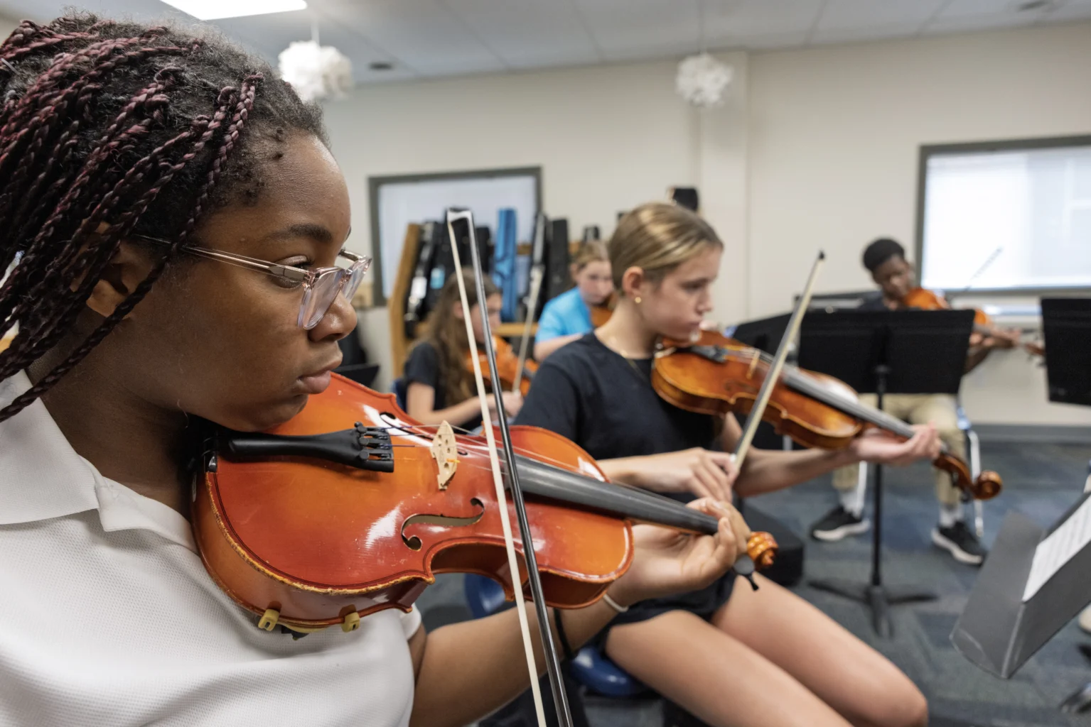 Students sit in a classroom playing violins, focusing on sheet music during a group practice session.