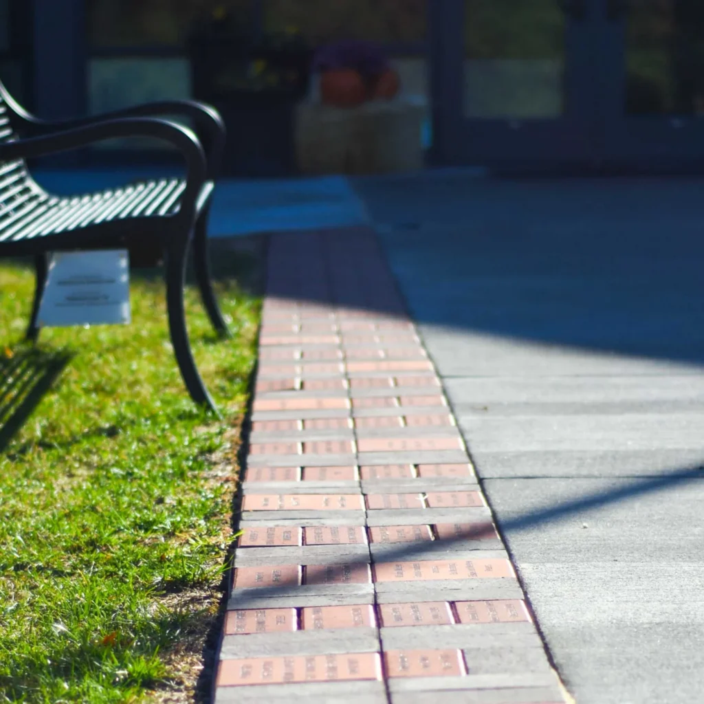 A row of engraved bricks forms a pathway beside a bench on grass, leading toward a building with glass doors in the background.