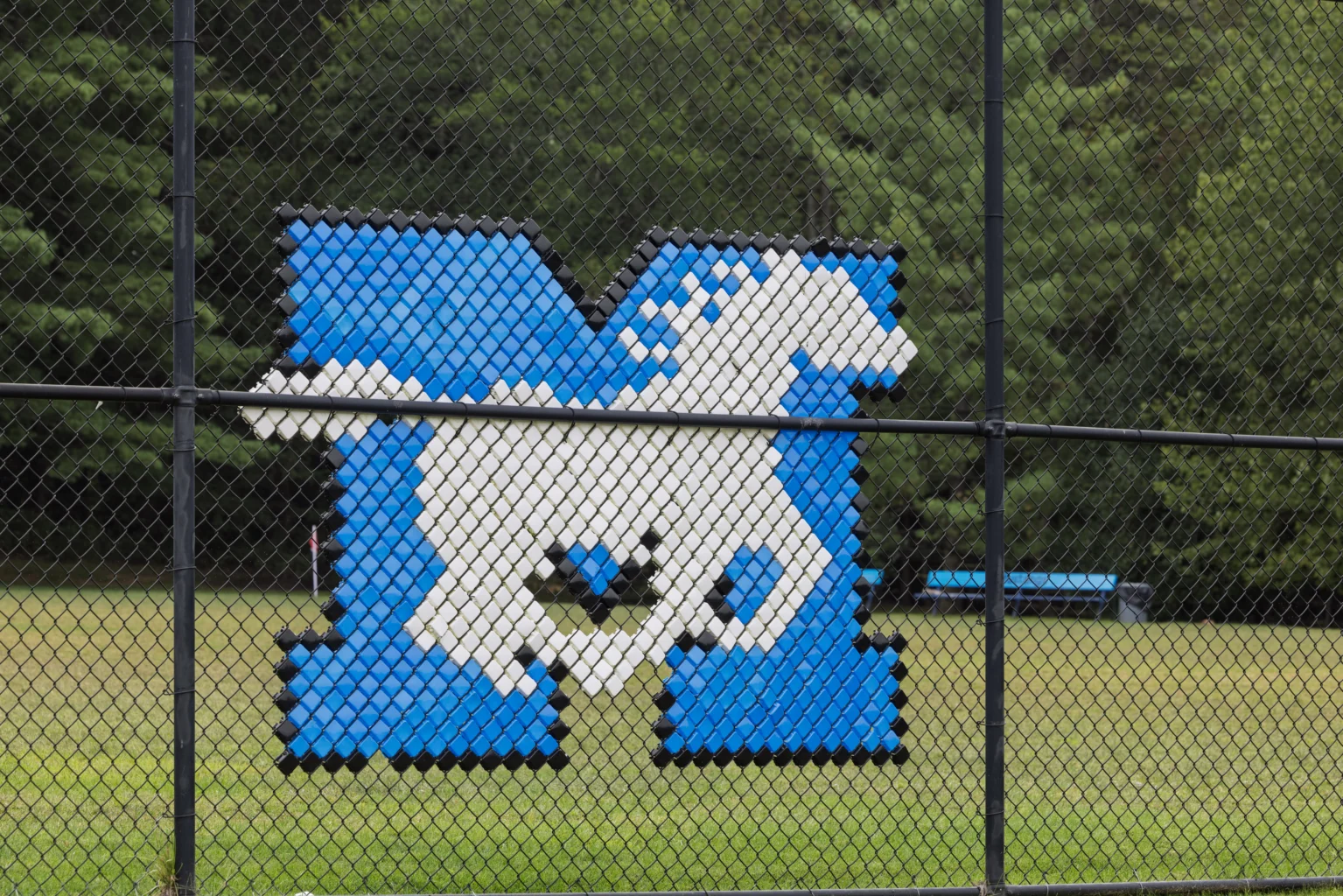 A blue letter "M" with a white horse design is displayed on a black chain-link fence, with trees and grass in the background.