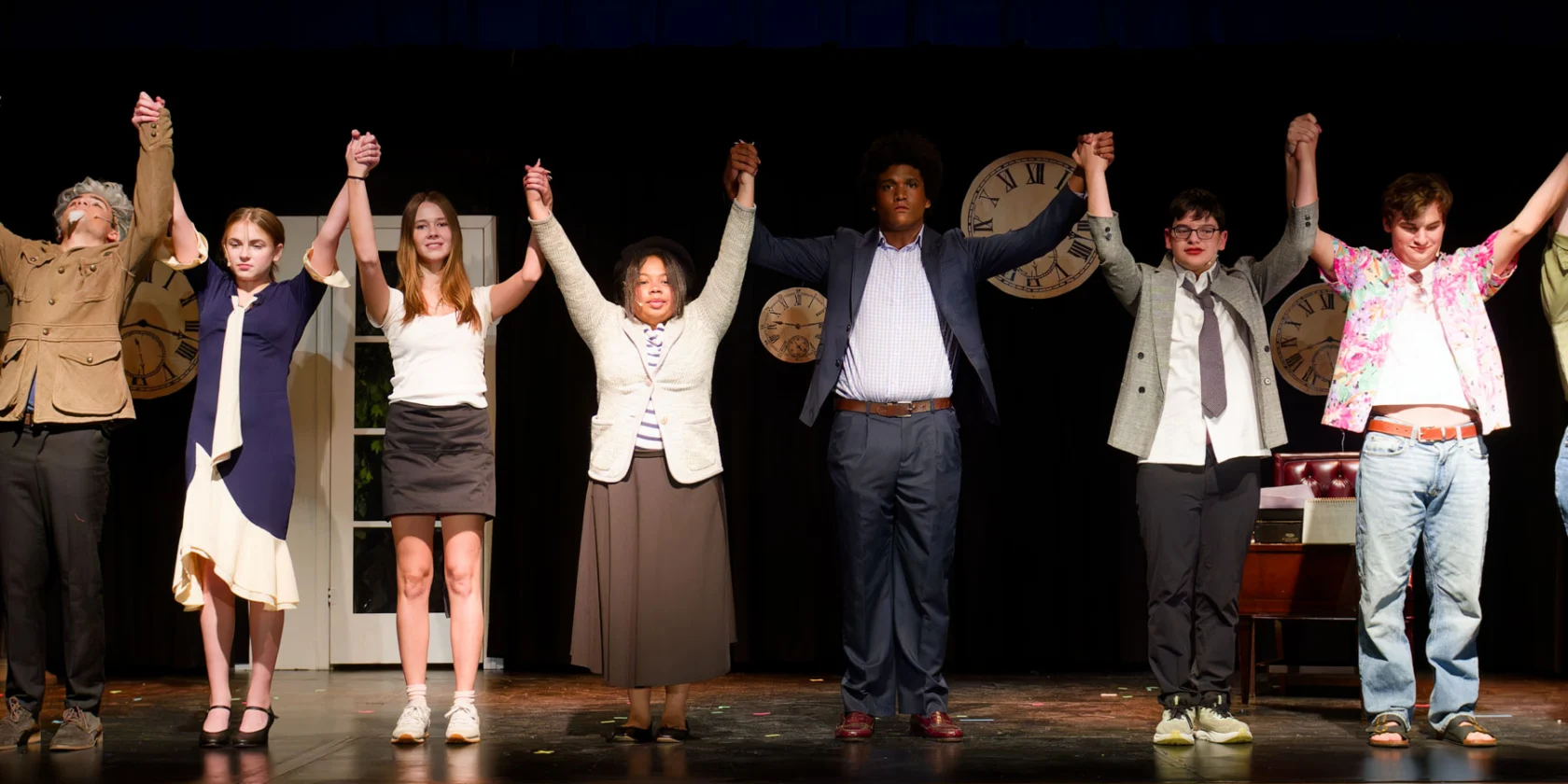 A group of actors in costumes stand on stage holding hands and raising their arms at the end of a theatrical performance.