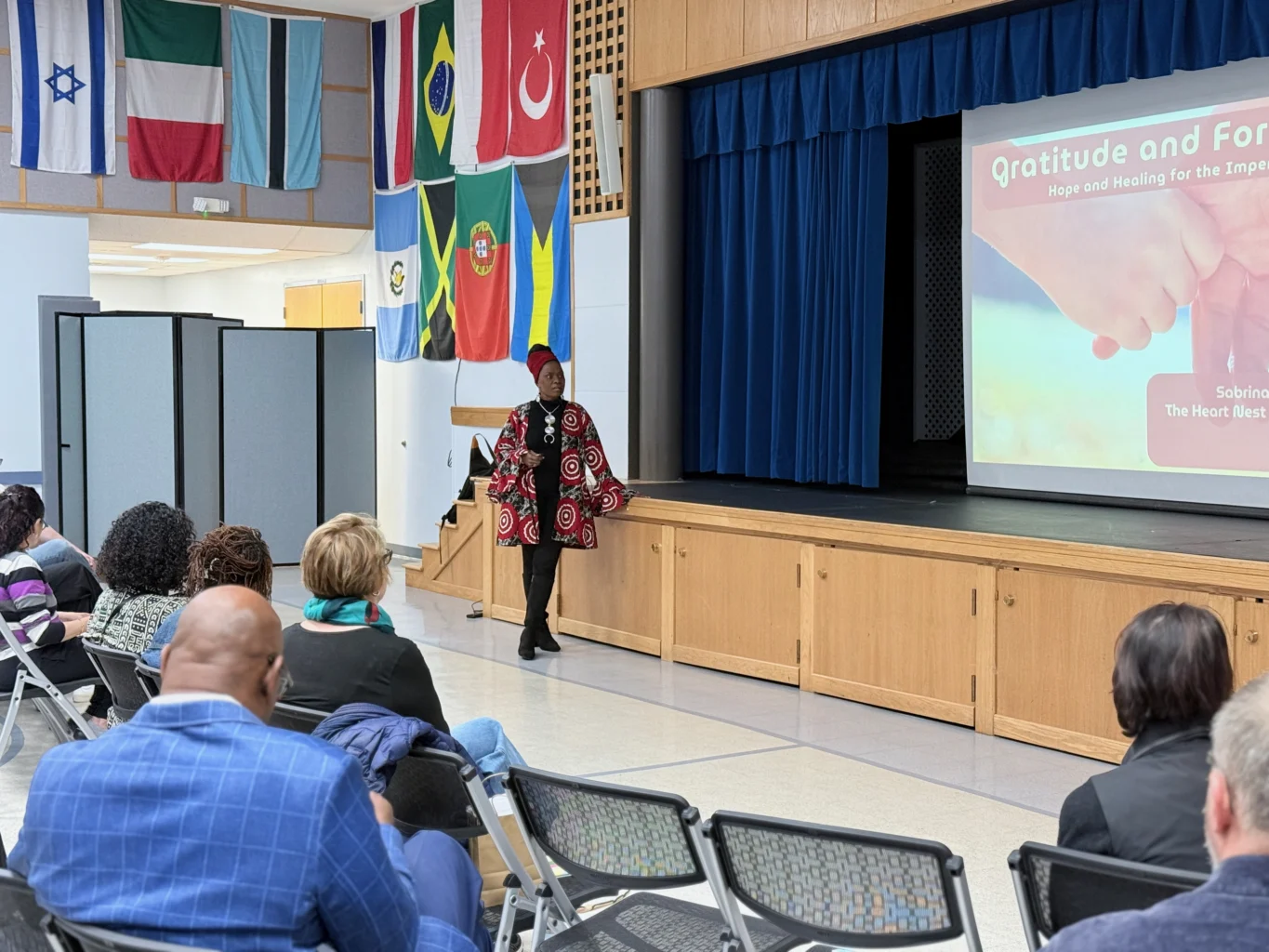 A speaker stands on stage in front of a projected presentation titled "Gratitude and Forgiveness" as an audience sits and listens in a room decorated with international flags.