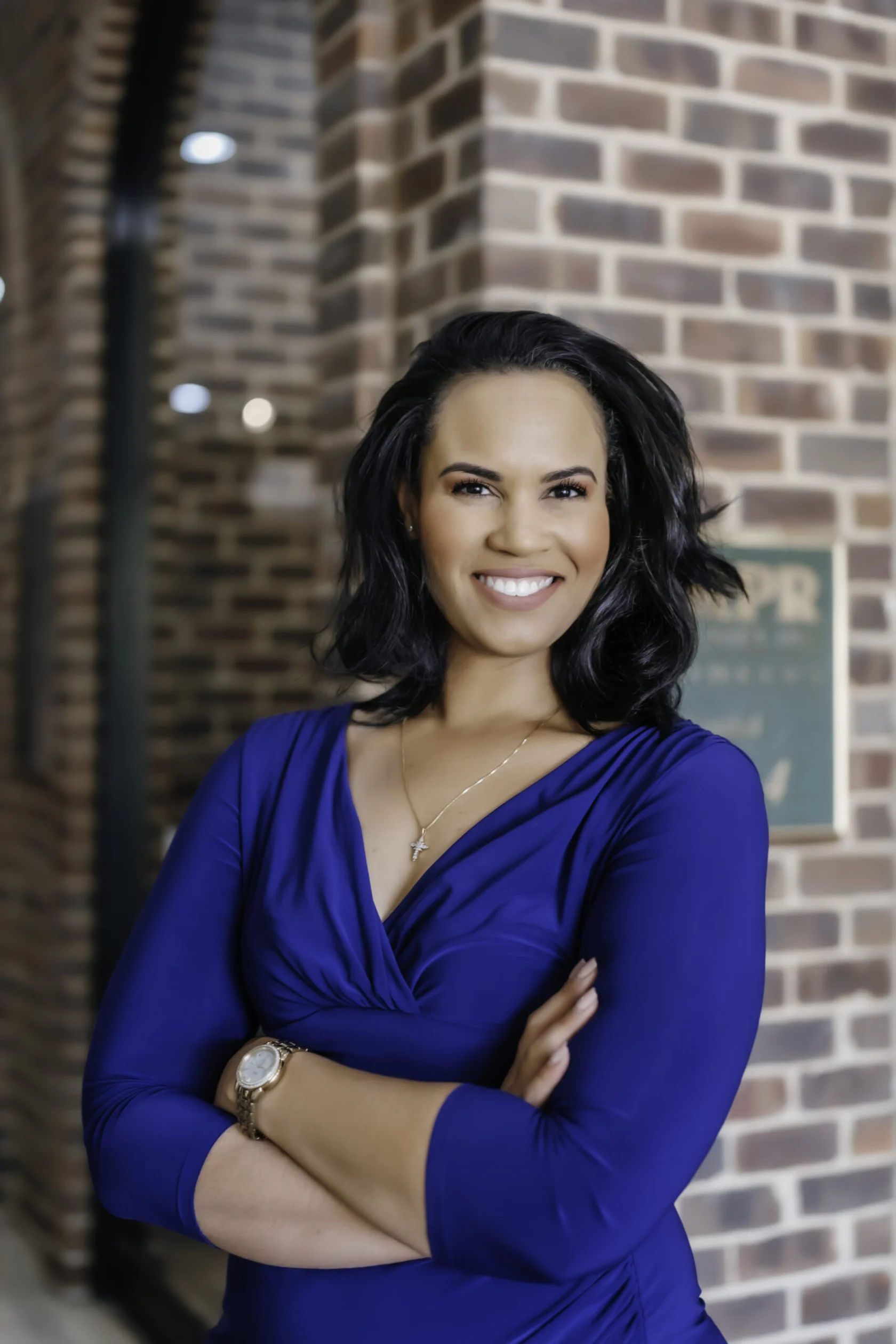 Woman with dark hair wearing a blue dress stands smiling with arms crossed in front of a brick wall.