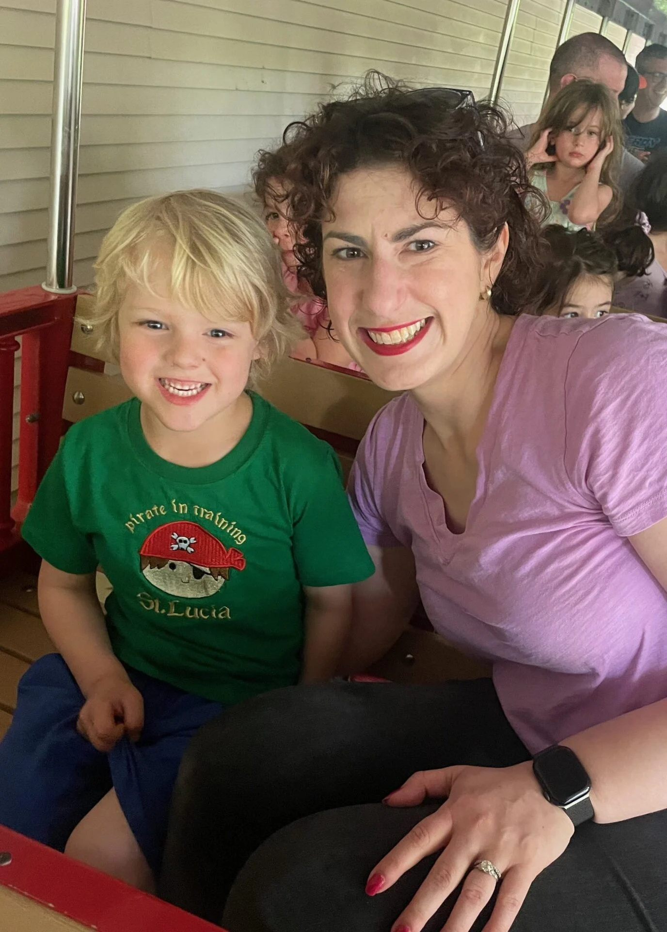 A woman and a young boy sit together and smile on a train ride, with other children and adults in the background.