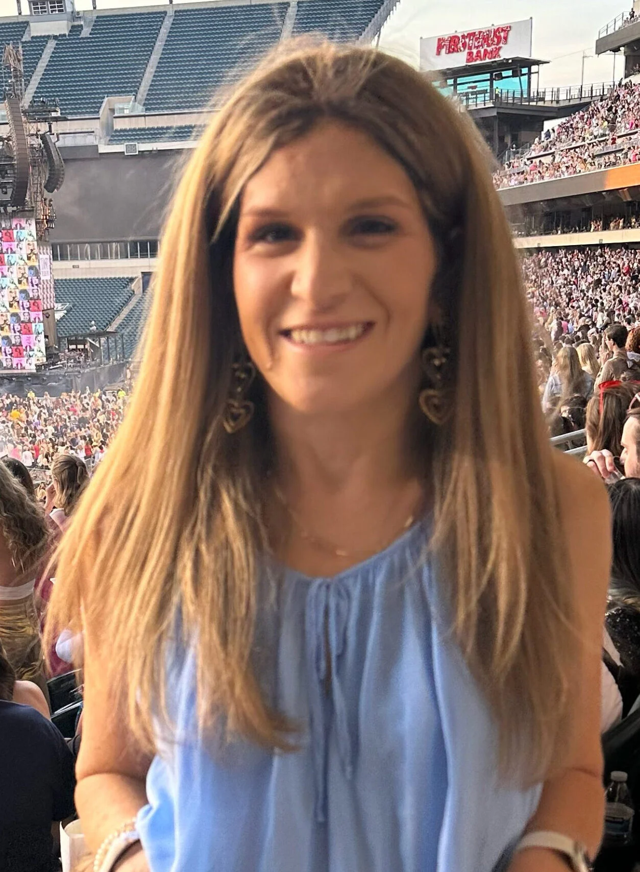 A woman with long light brown hair wearing a sleeveless blue top stands in a crowded stadium, smiling at the camera.