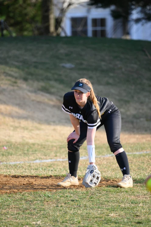 A softball player in black uniform and visor stands ready in an infield position, with hands on knees and a glove on her left hand, on a grassy field.