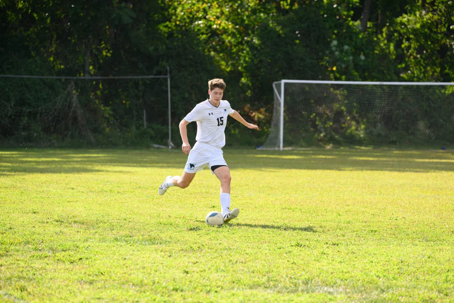 Soccer player in a white uniform with number 15 runs toward a ball on a grassy field, with a goal and trees in the background.