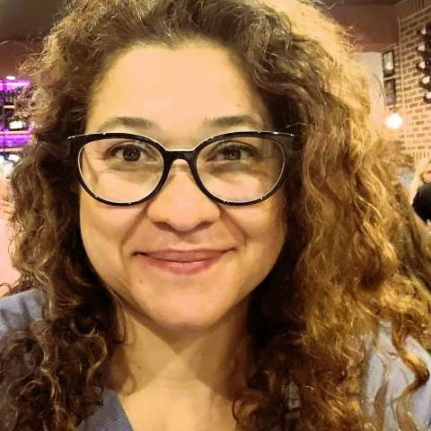 A person with curly brown hair and black glasses smiles at the camera in an indoor setting with warm lighting.