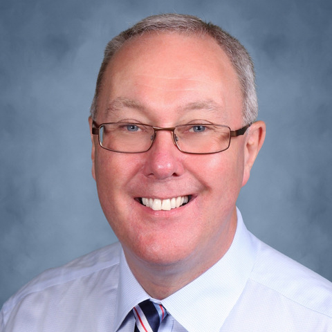 A middle-aged man with short gray hair and glasses, smiling, wearing a light blue shirt and striped tie, in front of a blue-gray background.