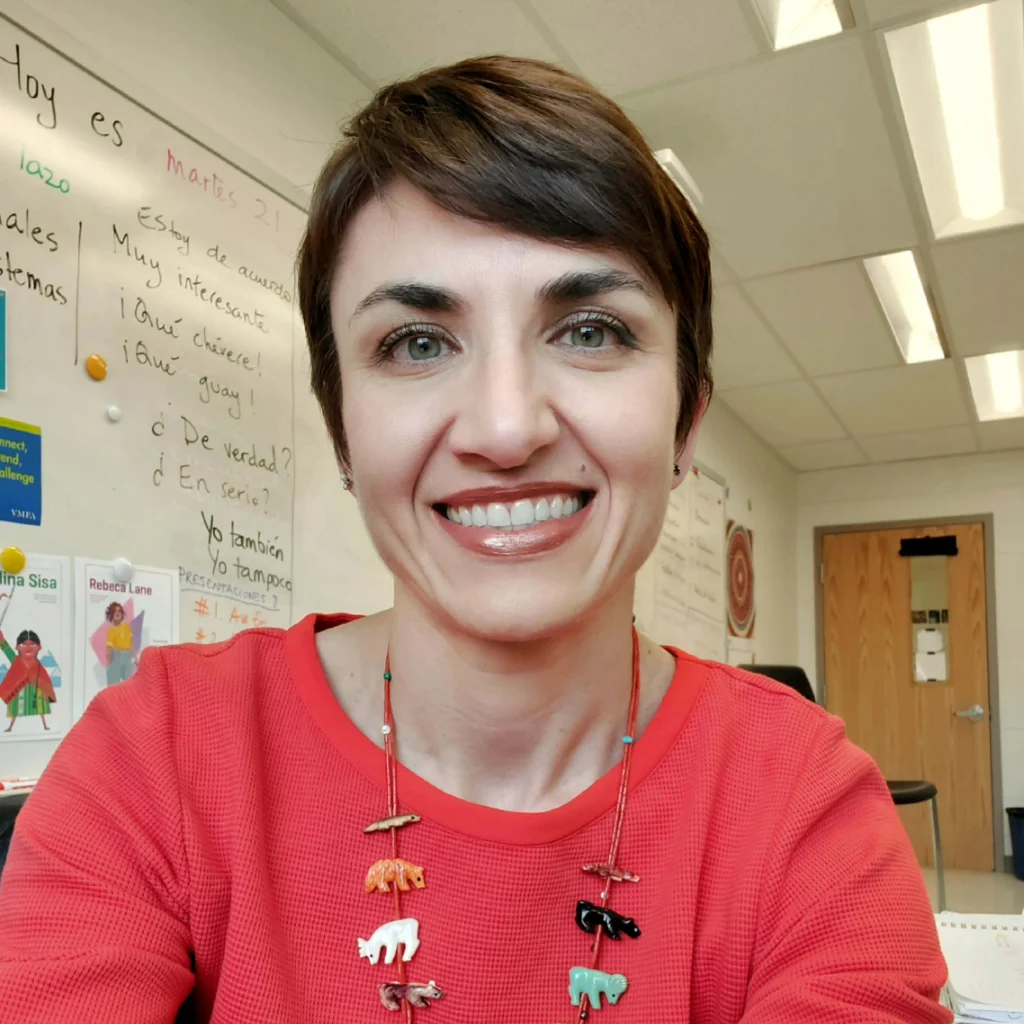 Woman with short brown hair wearing a red top and colorful animal necklace, smiling in a classroom with educational posters and a whiteboard in the background.