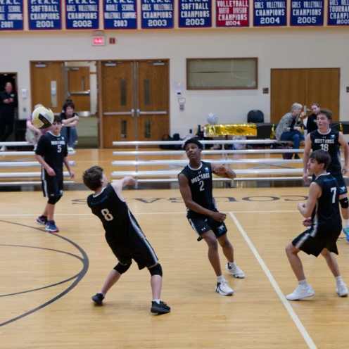 Five boys in black jerseys play volleyball in a gym, with one player about to hit the ball while others watch. Spectators sit on the bleachers in the background.