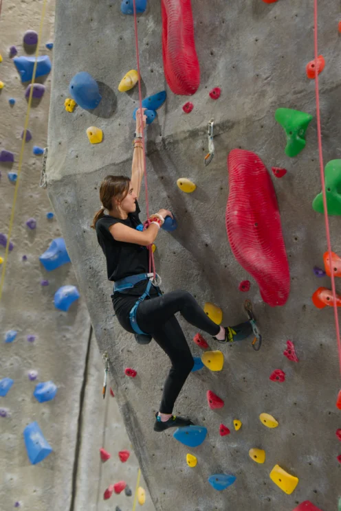 A person wearing a harness climbs an indoor rock wall with various colored holds, including large red, blue, and yellow grips.