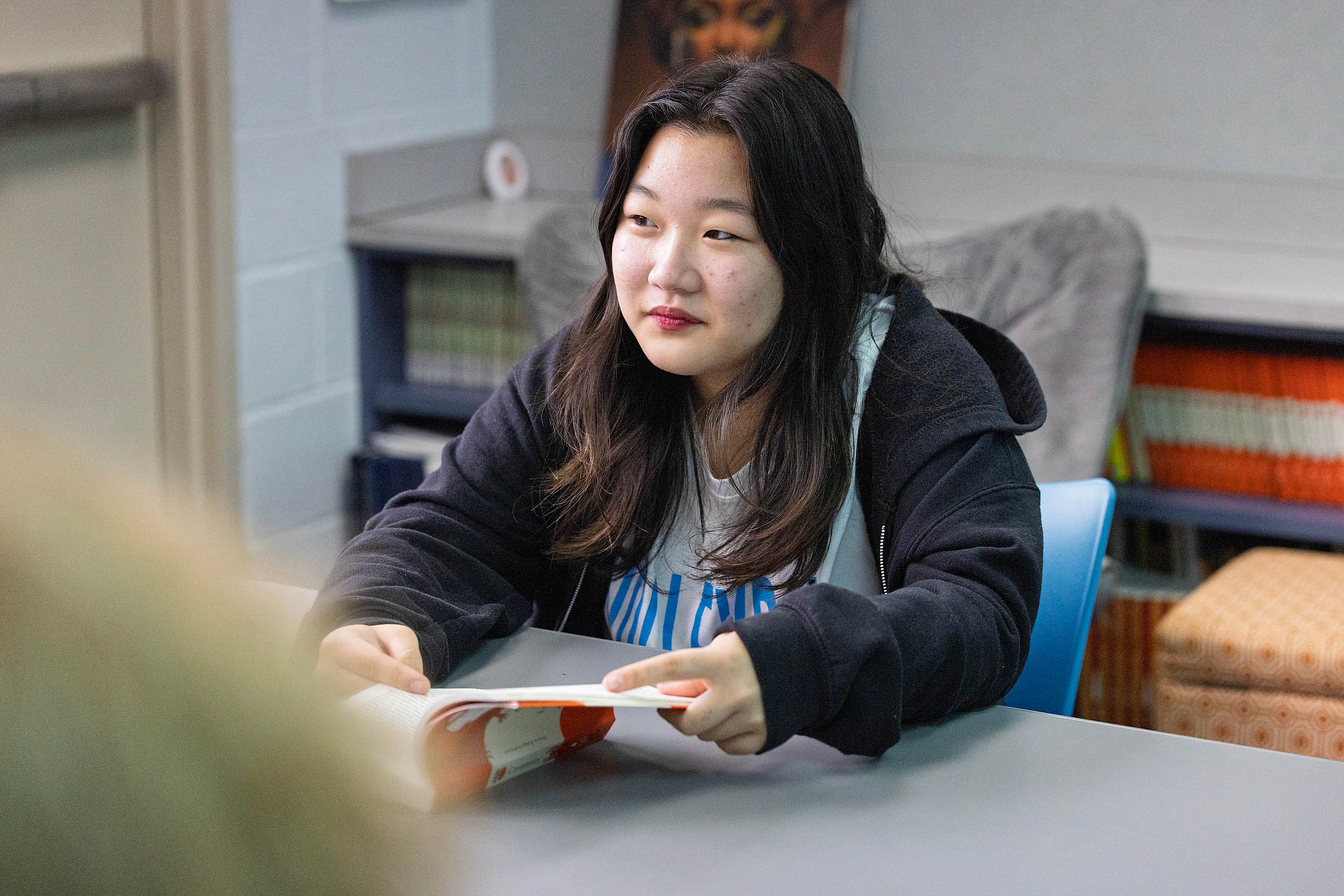 A young woman sits at a table holding an open book, looking slightly to the side. Shelves with books are visible in the background.