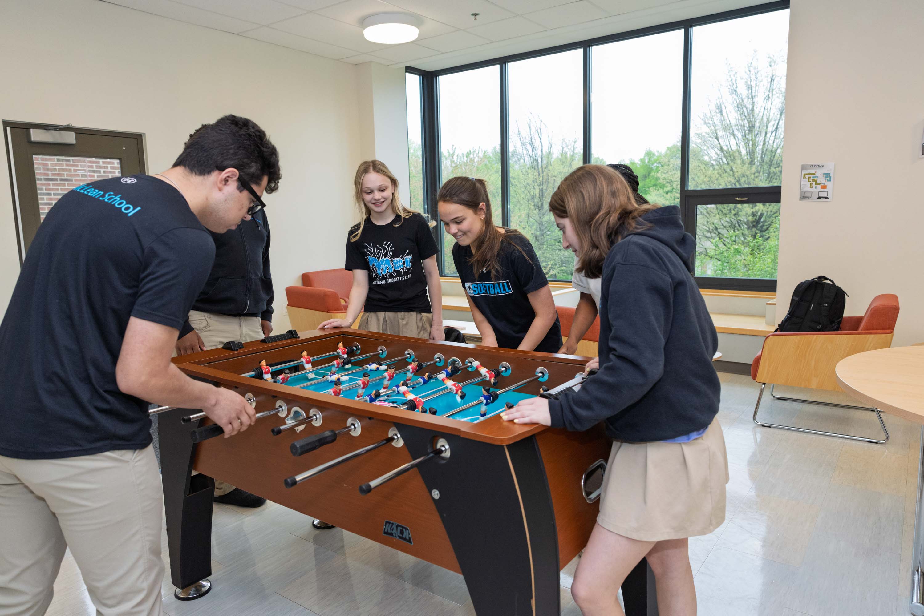 Four students play foosball together in a bright room with large windows, wearing school uniforms of navy tops and khaki bottoms.