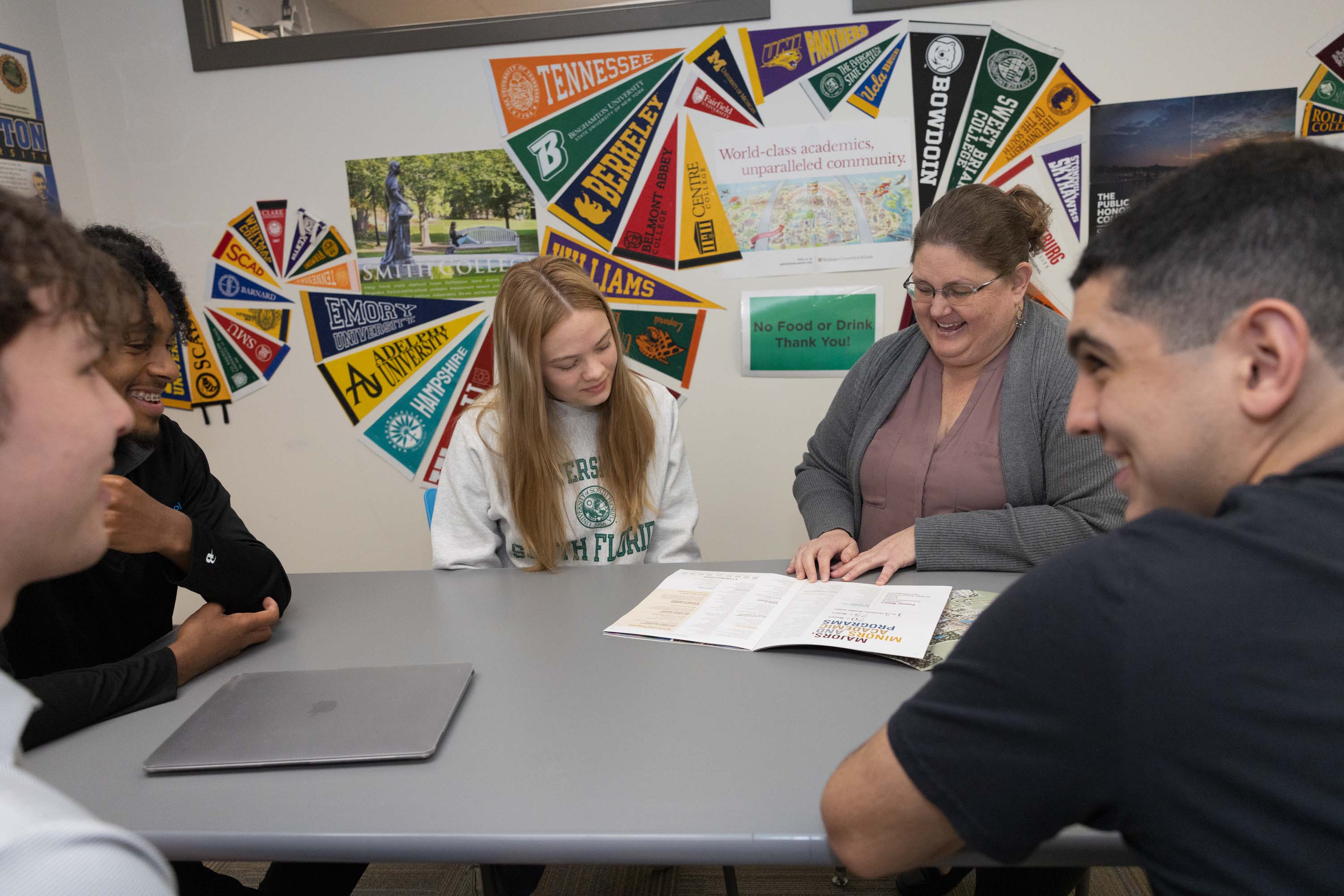 A group of four students and an adult sit around a table, looking at papers together, with college pennants displayed on the wall behind them.