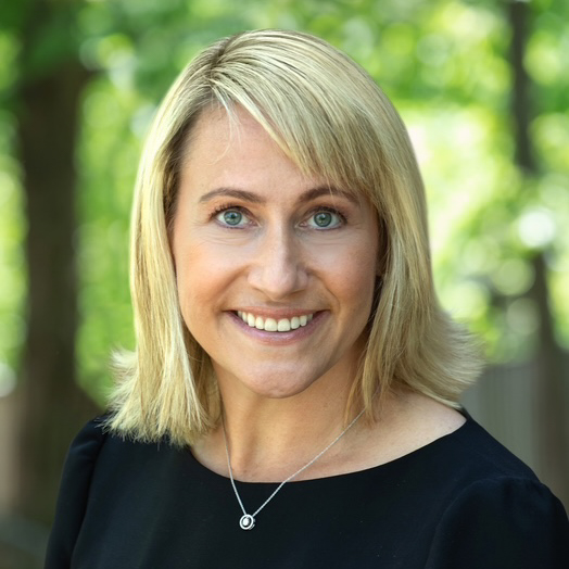 A woman with straight blonde hair and blue eyes smiles at the camera, wearing a black top and a silver necklace, with a blurred green outdoor background.