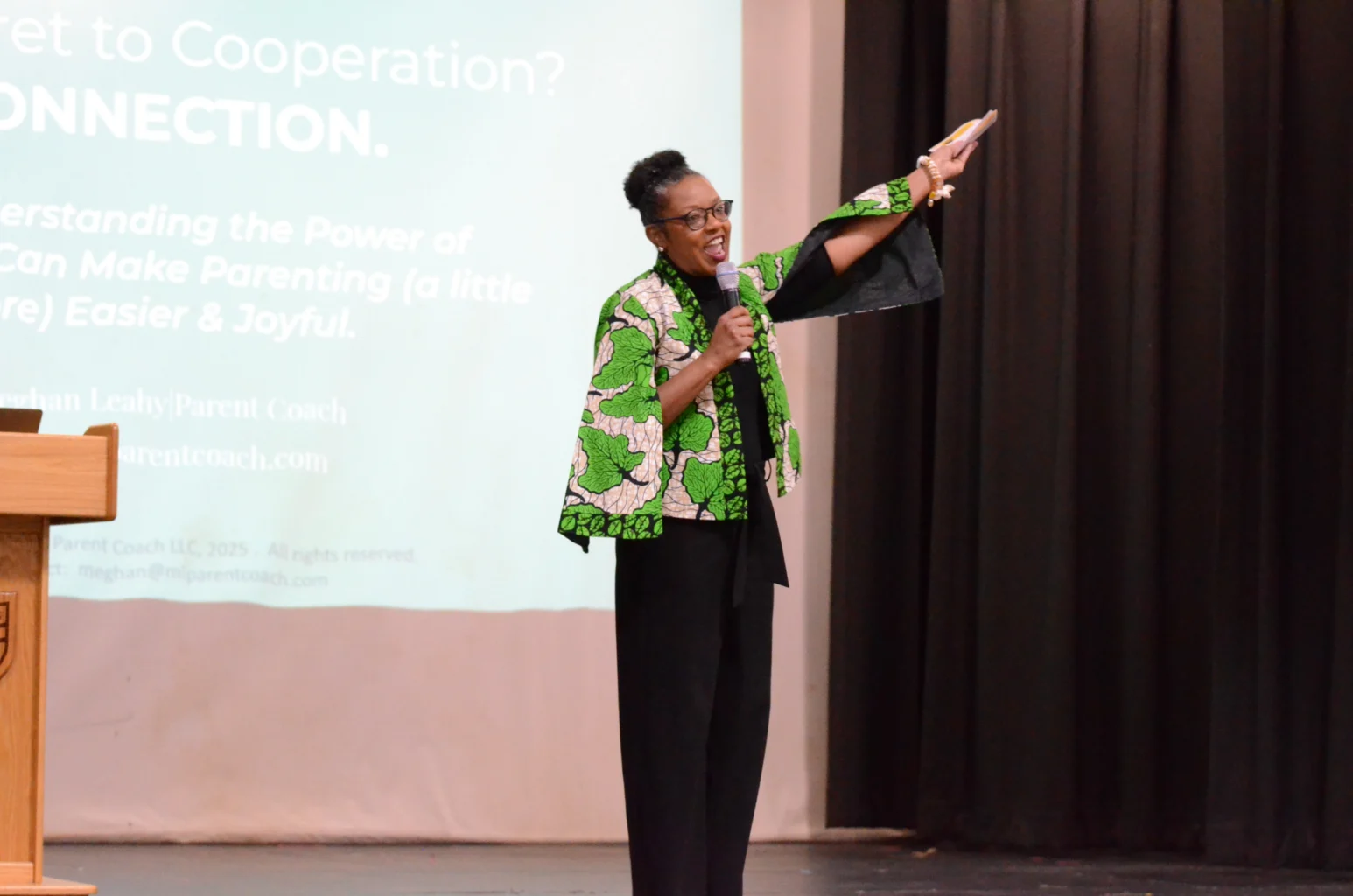 A woman stands on stage speaking into a microphone and gesturing with her right arm, with a presentation slide projected behind her.