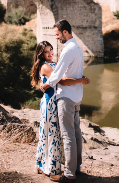 A couple stands outdoors by a river and historic stone bridge, embracing and smiling at the camera. The woman wears a white and blue dress; the man wears a white shirt and light pants.