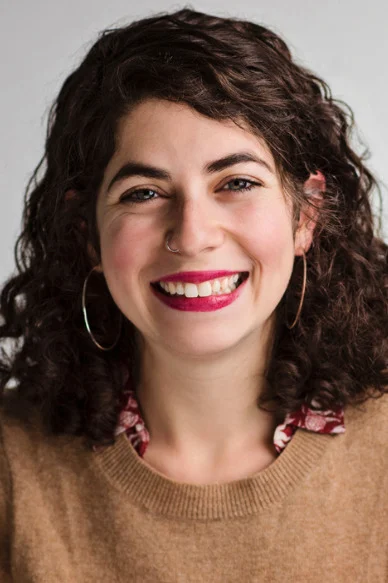 Woman with curly dark hair, wearing hoop earrings, a nose ring, a brown sweater, and a floral shirt, smiling at the camera against a neutral background.