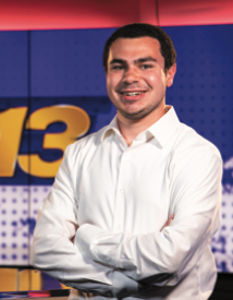 A person in a white dress shirt stands with arms crossed in front of a blue and yellow background with the number 13.