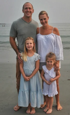 A man and woman stand behind two young girls on a beach, all barefoot and smiling at the camera with the sea in the background.