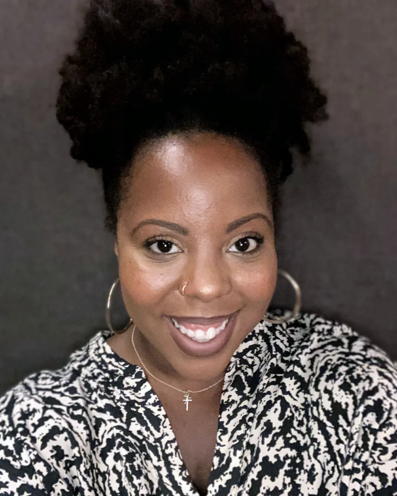 A woman with natural curly hair, hoop earrings, and a nose ring smiles at the camera, wearing a patterned top and a cross necklace against a plain dark background.