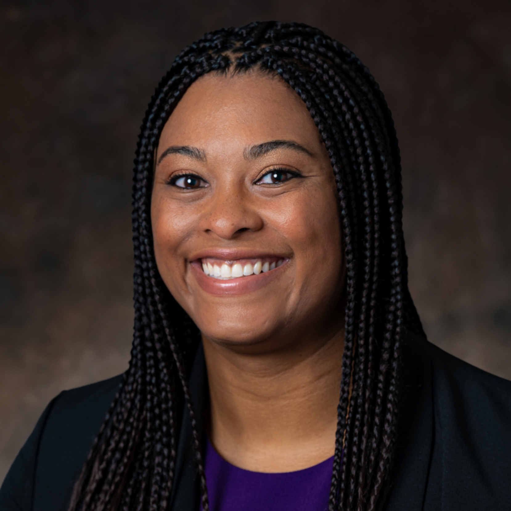 A woman with braided hair wearing a black blazer and purple top smiles at the camera against a dark background.