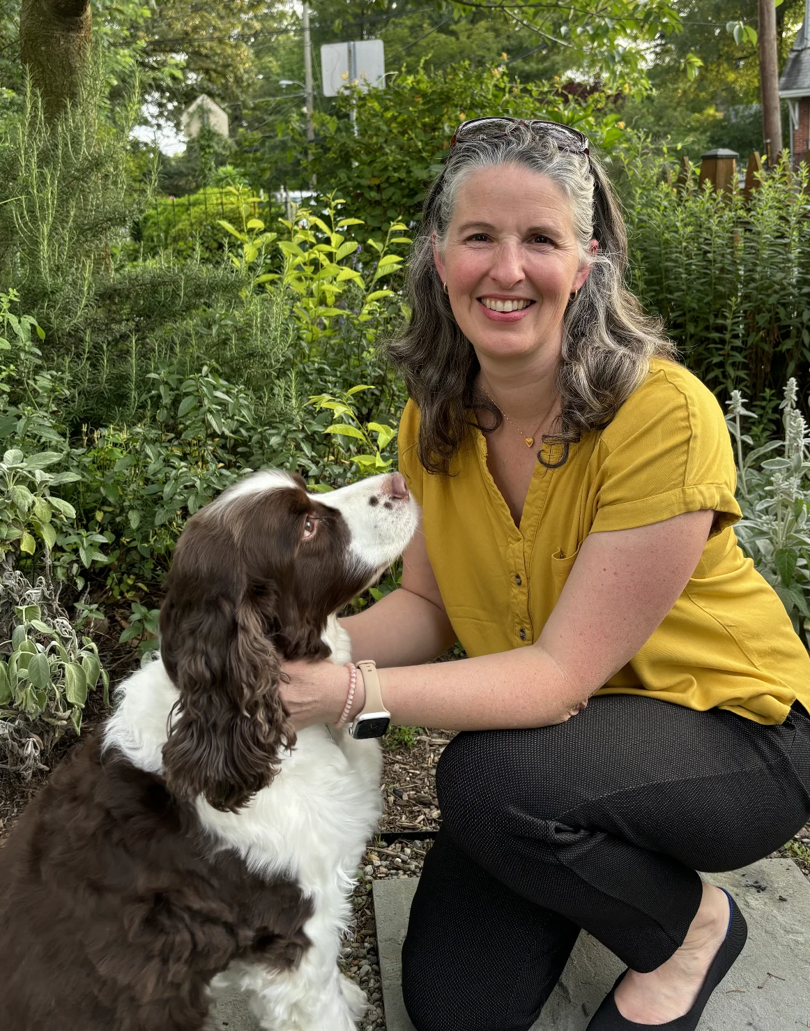 A woman in a yellow shirt kneels outdoors, smiling and petting a brown and white dog in a garden.