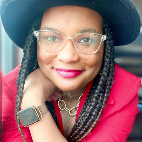 A person wearing glasses, a black hat, a red jacket, and a gold watch, smiling at the camera with their chin resting on their hand.