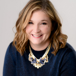 A woman with shoulder-length wavy blonde hair wearing a dark blue top and a large statement necklace, smiling at the camera.