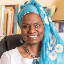 A woman wearing a blue headscarf with white patterns, blue jewelry, and a white top, sits and smiles in front of a bookshelf.