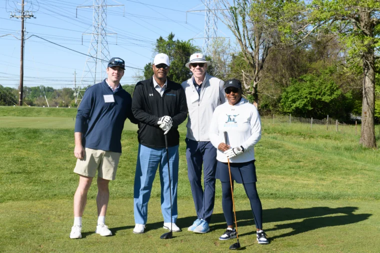 Four people wearing golf attire stand with clubs on a golf course, posing for a group photo. Power lines and trees are visible in the background.