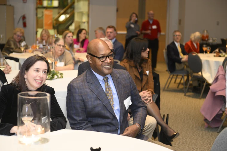 A group of people seated at round tables in a well-lit room, attentively watching an event or presentation.
