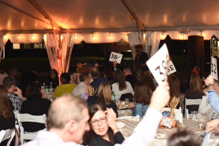 People seated at tables under a tent during an auction event, with several attendees holding up numbered bidding paddles.