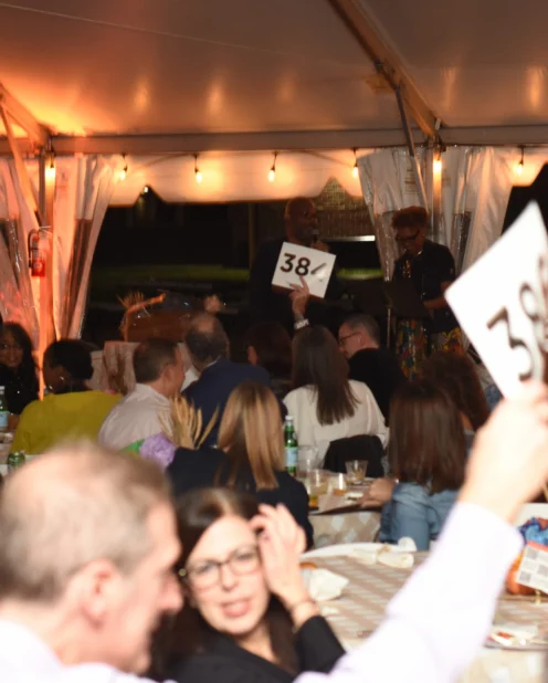 People seated at tables under a tent participate in an auction, with several individuals raising numbered paddles.