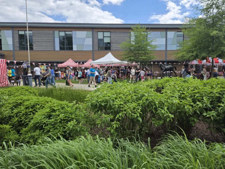 People gather at an outdoor event with red-and-white striped tents and bunting near a modern building, surrounded by greenery on a partly cloudy day.