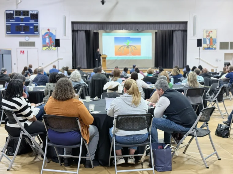 A group of people seated at round tables in a gymnasium watch a speaker presenting a slide on stage.