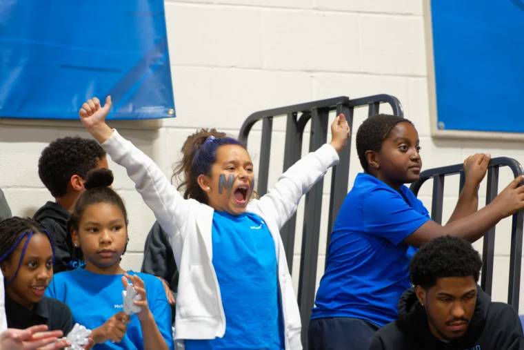 A young girl with face paint cheers with her arms raised among a group of children wearing blue shirts in a gym setting.