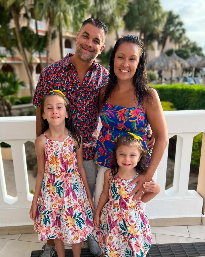 A man and woman stand behind two young girls, all wearing floral-patterned clothing, posing and smiling outdoors in front of a balcony with greenery and buildings in the background.