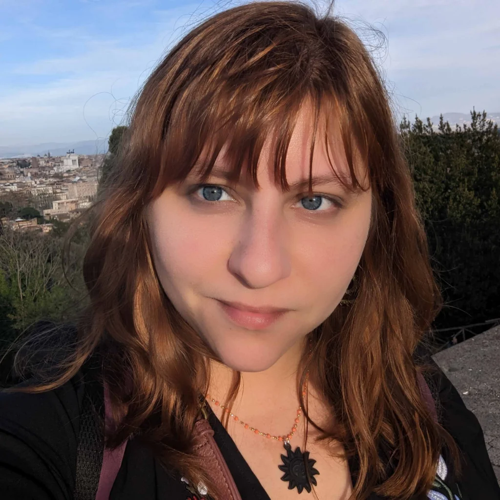 A person with light brown hair and blue eyes stands outdoors with a cityscape and trees in the background, wearing a black top and a sun-shaped necklace.
