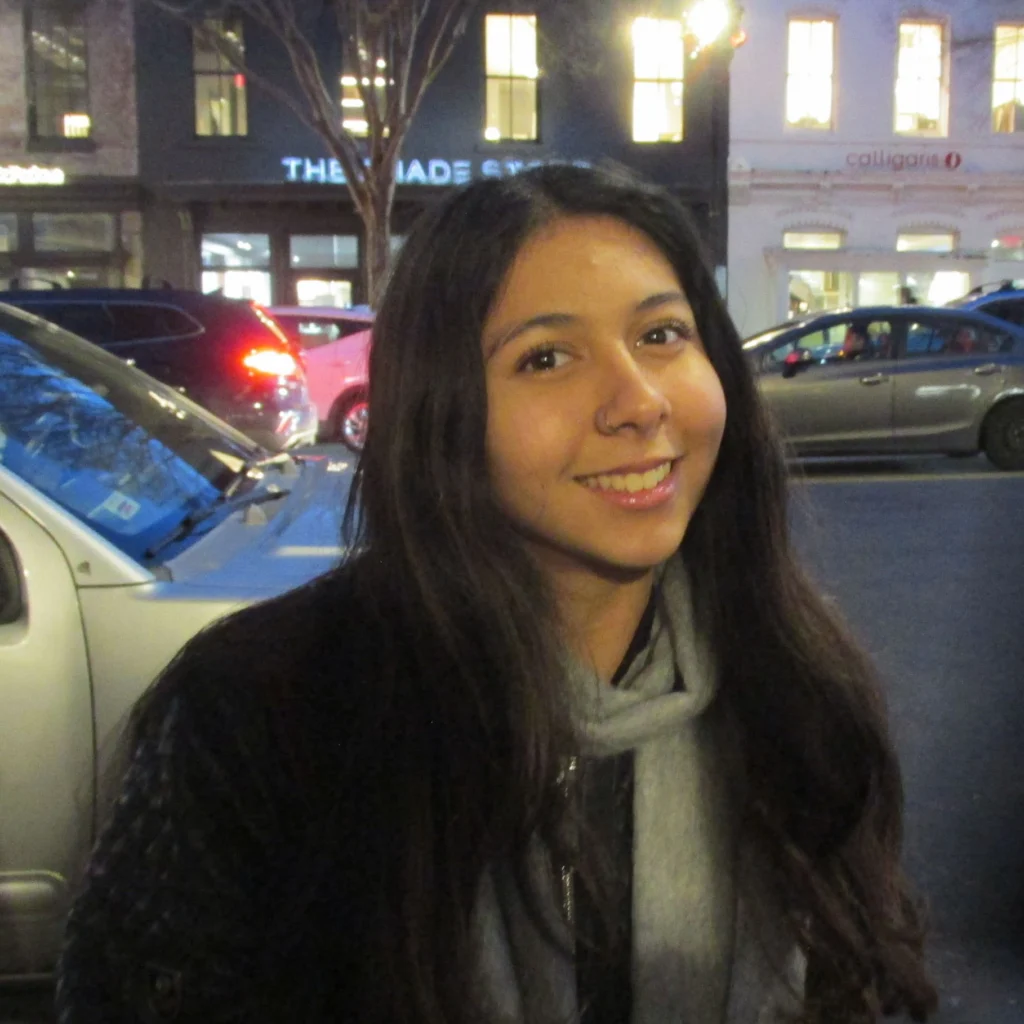 A young woman with long dark hair and a scarf smiles while sitting outdoors on a city street in the evening. Cars and buildings are visible in the background.